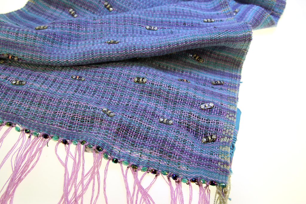 A hand-woven blue and violet textile with pink fringe and small beads