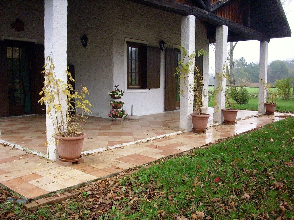 A white country house with a covered terracotta porch, potted plants and a green lawn